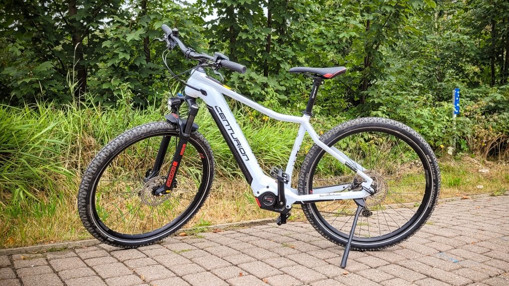 Weißes Mountainbike auf gepflastertem Weg vor Bäumen.