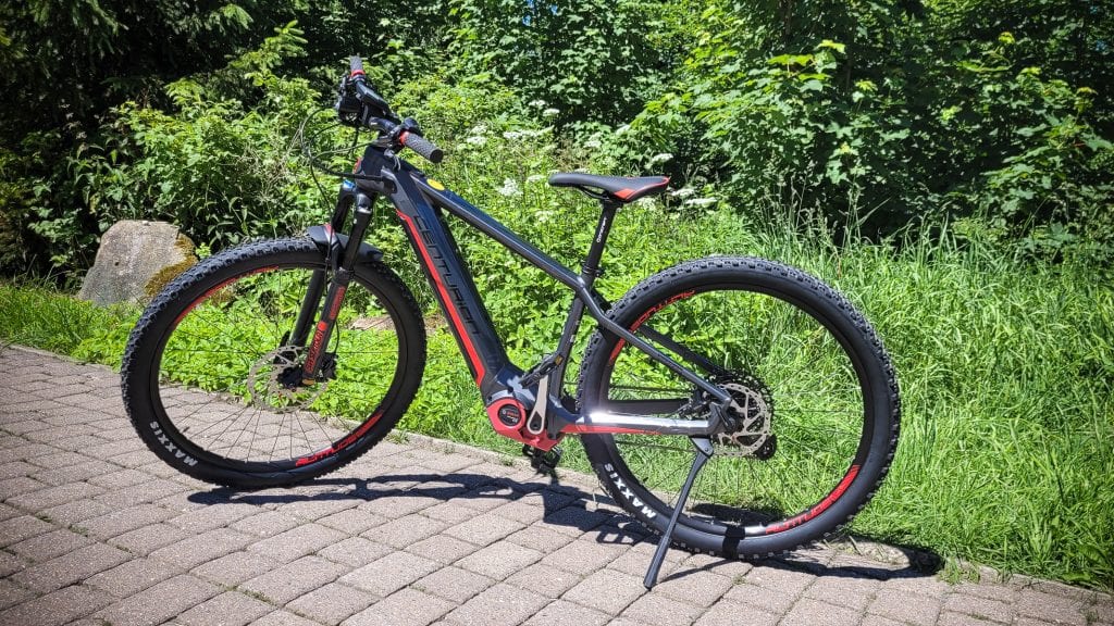 Schwarzes E-Bike auf Fahrradweg vor grüner Hecke.