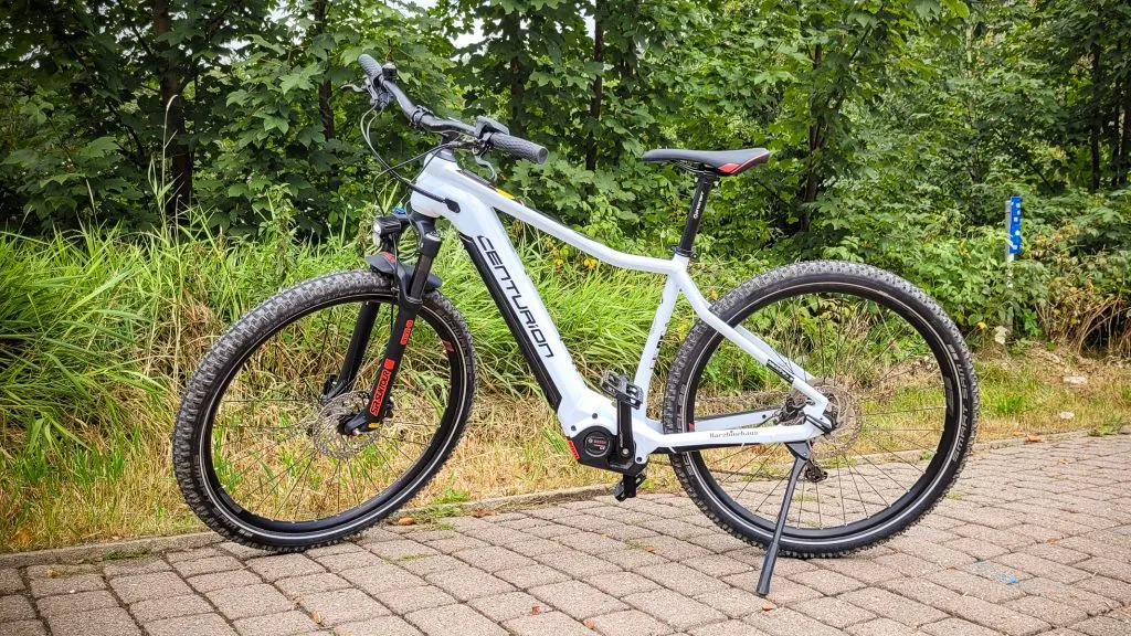 Weißes Mountainbike auf gepflastertem Weg vor Bäumen.